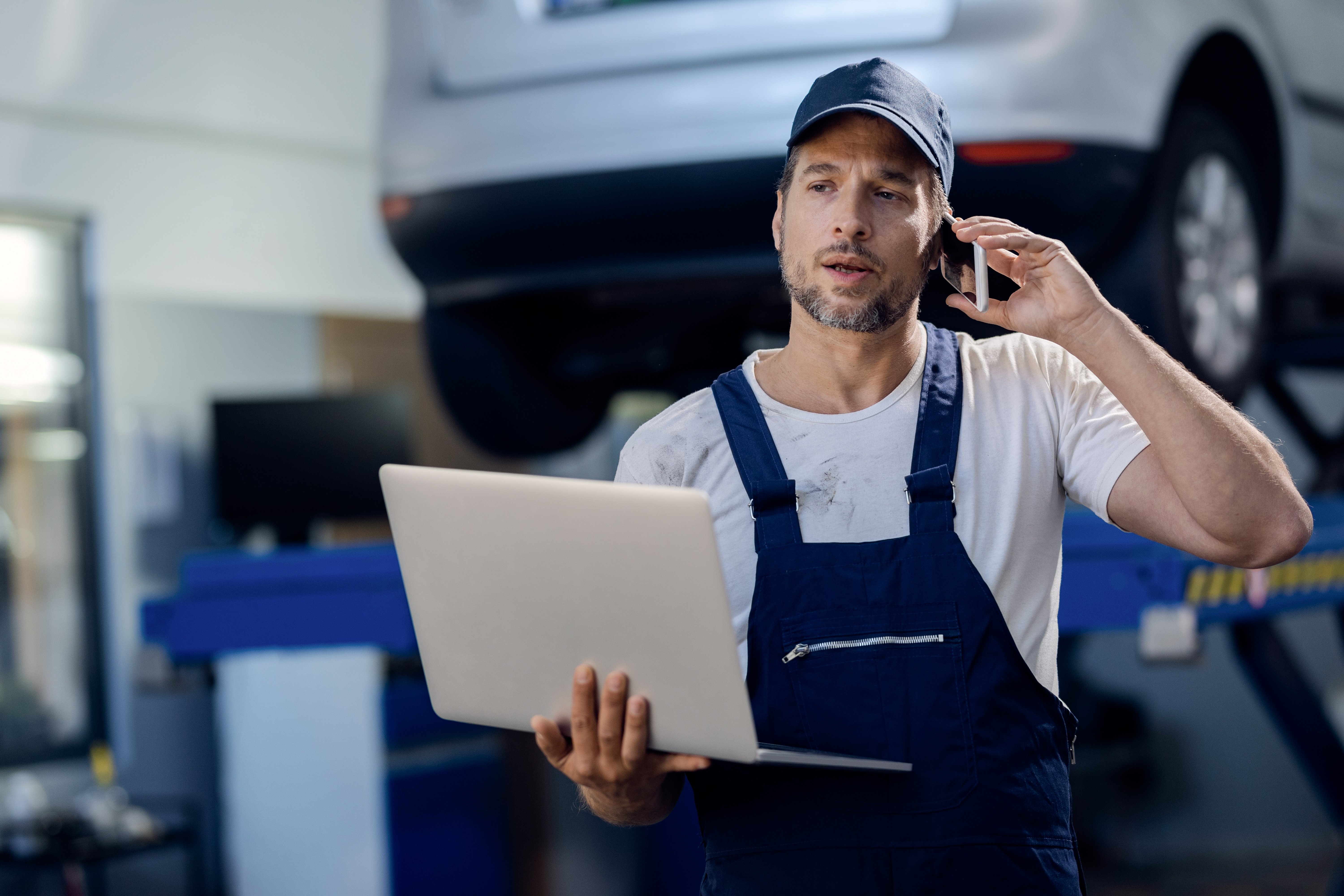 auto-repairman-talking-phone-while-using-laptop-workshop